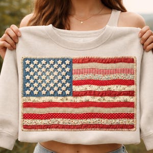 May include: A pale grey sweatshirt with a sewn-on American flag design. The flag features a blue denim field with white stars and red and white striped sections made of patterned fabric. The flag is framed with a decorative stitch.