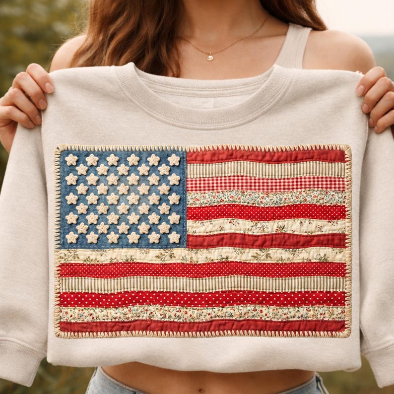 May include: A pale grey sweatshirt with a sewn-on American flag design. The flag features a blue denim field with white stars and red and white striped sections made of patterned fabric. The flag is framed with a decorative stitch.