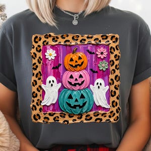 Puede incluir: Una camiseta gris carb&oacute;n con un gr&aacute;fico cuadrado con borde de estampado de leopardo. El centro muestra una pila de tres calabazas de Halloween en naranja, rosa y turquesa, flanqueadas por dos fantasmas blancos, flores y murci&eacute;lagos sobre un fondo morado.