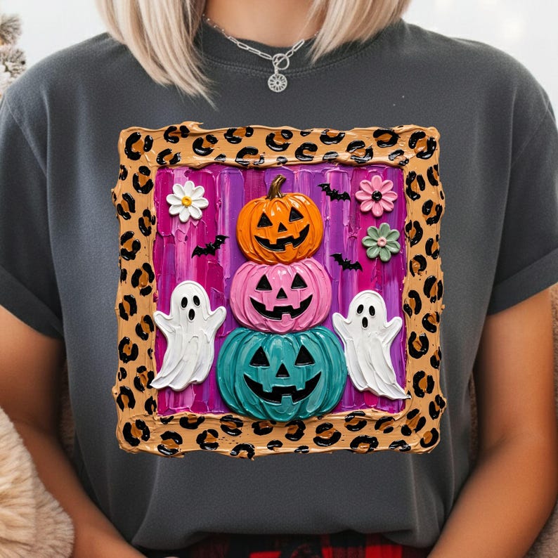 Puede incluir: Una camiseta gris carb&oacute;n con un gr&aacute;fico cuadrado con borde de estampado de leopardo. El centro muestra una pila de tres calabazas de Halloween en naranja, rosa y turquesa, flanqueadas por dos fantasmas blancos, flores y murci&eacute;lagos sobre un fondo morado.