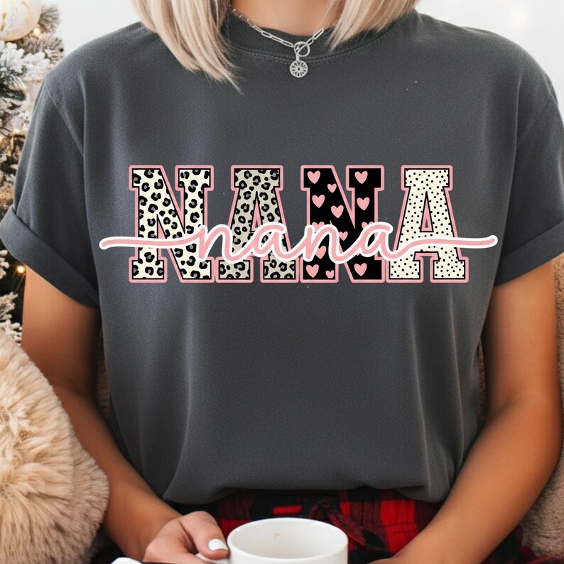 May include: Dark gray t-shirt with the word "NANA" in a decorative font. The letters are filled with leopard print, hearts, and polka dots, outlined in pink. The word "nana" is written in cursive below the letters.