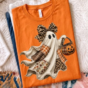 Pu&ograve; includere: Una t-shirt arancione a girocollo con la grafica di un fantasma composto da vari ritagli di tessuto. Il fantasma indossa un fiocco leopardato e tiene in mano un secchiello a forma di zucca a quadri marroni. Il design include motivi floreali e animalier.