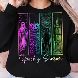 May include: A black sweatshirt featuring a graphic print with four vertical panels in neon colors. The panels depict a ghost with a sunflower, a pumpkin on books with sunflowers, a black cat with a spiderweb, and a gothic church. Text reads Spooky Season.