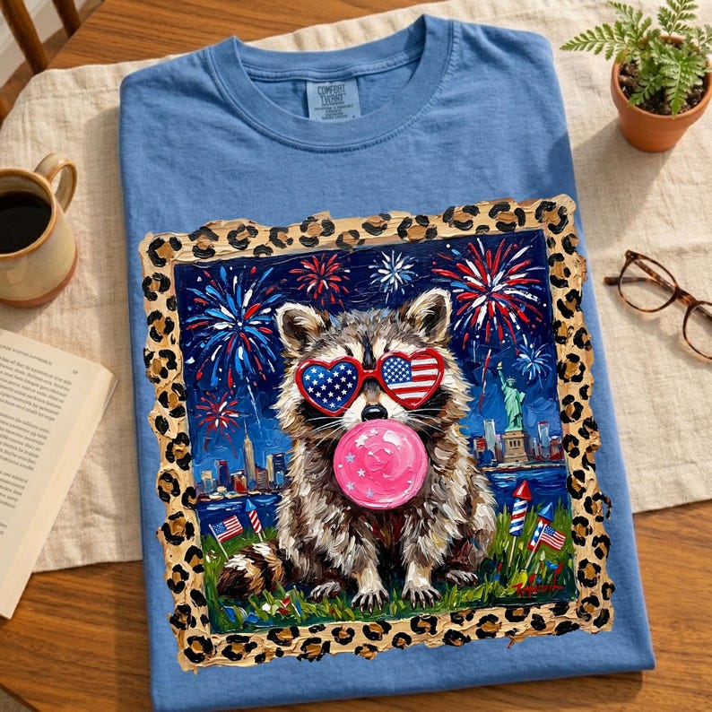 May include: A light blue t-shirt featuring a raccoon wearing heart-shaped sunglasses with the American flag design, blowing a pink bubble. The raccoon is set against a backdrop of fireworks, the Statue of Liberty, and a cityscape. The image is framed with a leopard print border.