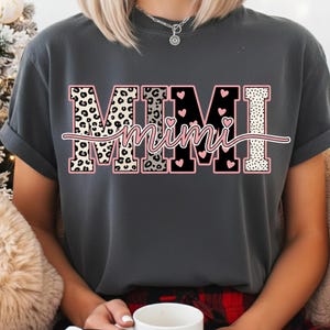 May include: Dark gray t-shirt with the word "MIMI" in a bold, decorative font. The letters have leopard print, black, and dotted patterns, with pink accents and hearts. The word "Mimi" is written in a cursive font across the letters.