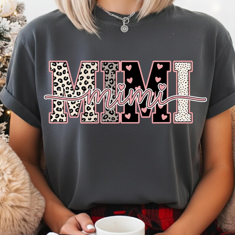 May include: Dark gray t-shirt with the word "MIMI" in a bold, decorative font. The letters have leopard print, black, and dotted patterns, with pink accents and hearts. The word "Mimi" is written in a cursive font across the letters.