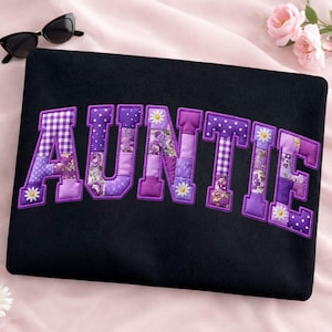 May include: Black tote bag with the word "AUNTIE" appliquéd in purple patchwork letters. The letters feature various patterns, including polka dots, floral prints, and gingham. Small white daisy accents are placed on the letters. Sunglasses are in the upper left corner.