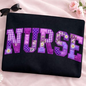 Könnte beinhalten: Schwarze Laptoptasche mit dem Wort "NURSE" aus lila Patchworkstoff. Die Buchstaben weisen verschiedene Muster auf, darunter Vichy, Tupfen, Blumendrucke und unifarbene Farben, mit Gänseblümchen-Akzenten. Sonnenbrille liegt auf der rosa Oberfläche.