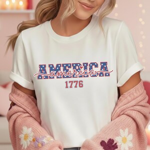 May include: White t-shirt with the word "America" in blue and red block letters with stars and hearts, and the year "1776" in red. The shirt has rolled-up sleeves.