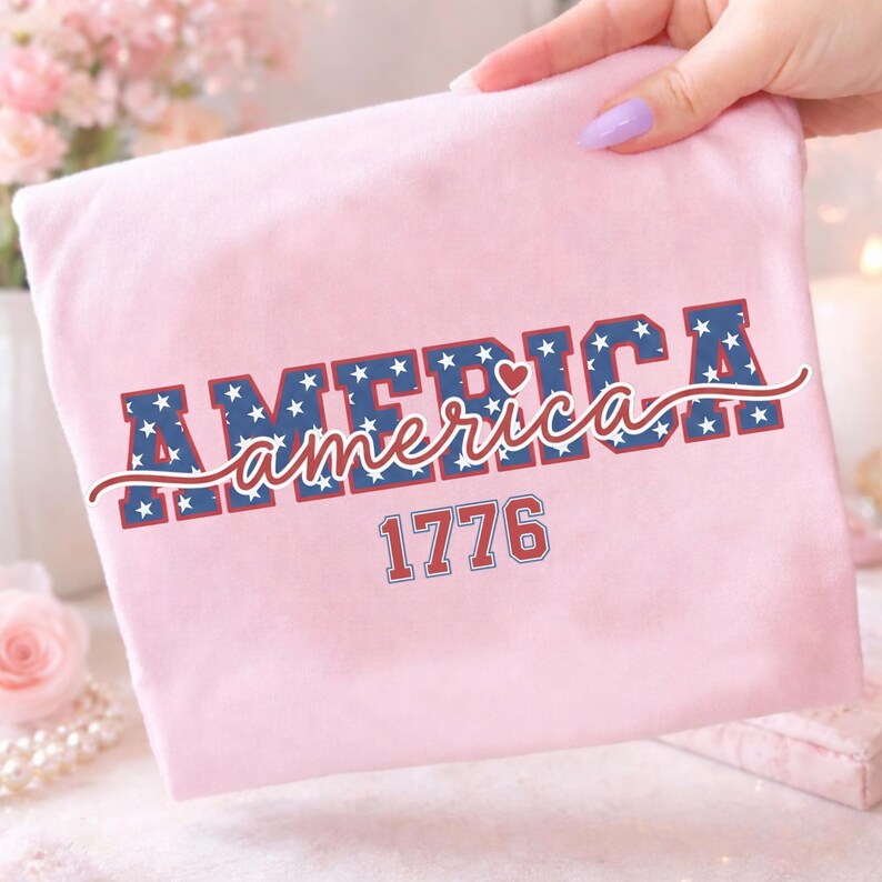 May include: A light pink t-shirt with the word "AMERICA" in a bold, blue font filled with white stars. The word "America" is written in a cursive font in red. The number "1776" is printed below in a red font.