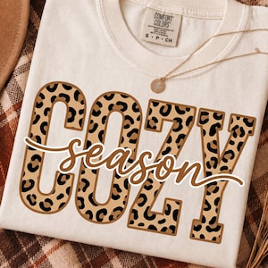 May include: An off-white short-sleeve t-shirt featuring the word COZY in large block letters with a brown and black leopard print pattern. The word season is written in a brown cursive script across the center. A gold necklace rests on the shirt.