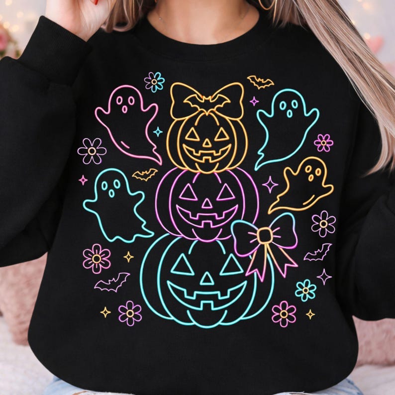 May include: A black long-sleeve sweatshirt featuring a neon-style graphic print of three stacked jack-o'-lanterns, ghosts, bats, and flowers. The design uses vibrant outlines in pink, yellow, and blue, with bows on the top and bottom pumpkins.