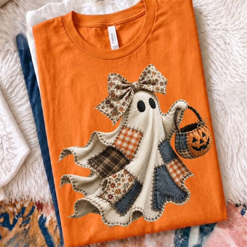 Op de afbeelding: Een oranje t-shirt met een afbeelding van een geest gemaakt van verschillende stoffen lapjes, waaronder ruitjes- en bloemenpatronen. De geest draagt een grote bijpassende stoffen strik op het hoofd en draagt een klein pompoenvormig mandje.