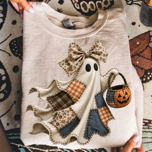 Op de afbeelding: Een beige sweater met ronde hals en een centrale afbeelding van een geest met een bloemrijke strik. De geest is versierd met diverse stoffen patches in oranje gingham, bruin ruitpatroon en bloemmotieven, en draagt een pompoenemmertje.
