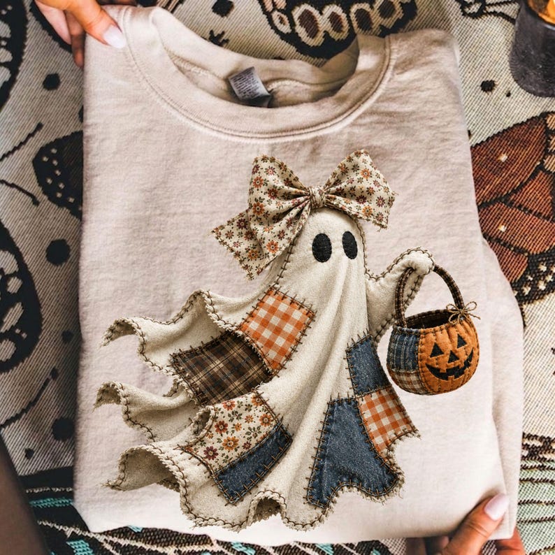 Op de afbeelding: Een beige sweater met ronde hals en een centrale afbeelding van een geest met een bloemrijke strik. De geest is versierd met diverse stoffen patches in oranje gingham, bruin ruitpatroon en bloemmotieven, en draagt een pompoenemmertje.