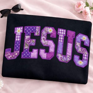 May include: Black fabric with the word "JESUS" appliqued in purple patchwork letters. The letters feature floral, polka dot, and gingham patterns, with daisy accents. Sunglasses are in the upper left corner.