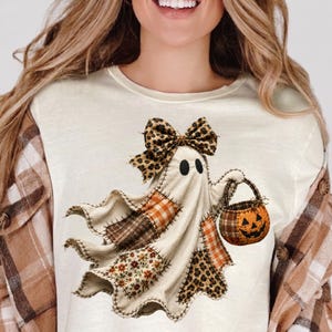 Pu&ograve; includere: Una t-shirt color crema con la grafica di un fantasma composto da vari ritagli di tessuto, tra cui quadri arancioni, fiori marroni e stampa leopardata. Il fantasma indossa un fiocco leopardato e tiene in mano un secchiello a zucca.