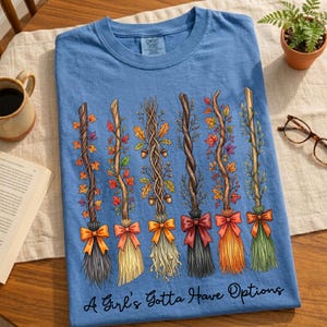 May include: A light blue t-shirt featuring a design of six decorative brooms with colorful bows and autumn foliage. The text "A Girl's Gotta Have Options" is printed at the bottom of the shirt. The shirt is displayed on a wooden surface.