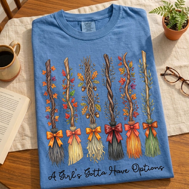 May include: A light blue t-shirt featuring a design of six decorative brooms with colorful bows and autumn foliage. The text "A Girl's Gotta Have Options" is printed at the bottom of the shirt. The shirt is displayed on a wooden surface.