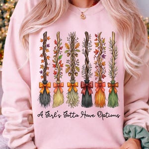 May include: Pink sweatshirt featuring six decorative broom designs with autumn leaves and bows. The text "A Girl's Gotta Have Options" is printed below the brooms. The brooms have different colors and styles.
