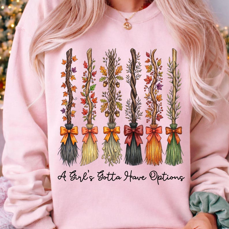 May include: Pink sweatshirt featuring six decorative broom designs with autumn leaves and bows. The text "A Girl's Gotta Have Options" is printed below the brooms. The brooms have different colors and styles.