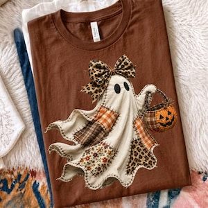 Pu&ograve; includere: Una t-shirt marrone a girocollo con la grafica di un fantasma composto da vari tessuti a motivi, tra cui tartan, fiori e stampa leopardata. Il fantasma indossa un grande fiocco leopardato e tiene in mano un piccolo secchiello a forma di zucca.