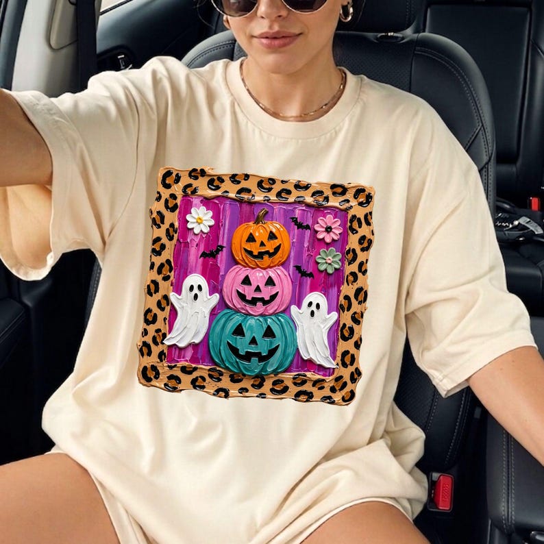 Puede incluir: Una camiseta de color crema con un estampado gr&aacute;fico cuadrado. El dise&ntilde;o incluye una pila de tres calabazas talladas en naranja, rosa y verde azulado, flanqueadas por dos fantasmas blancos, peque&ntilde;os murci&eacute;lagos y flores, todo enmarcado por un borde de estampado de leopardo marr&oacute;n.