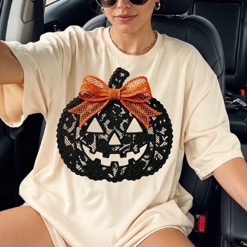 Może przedstawiać: Kremowy t-shirt z kr&oacute;tkim rękawem z dużą grafiką dyni wykonanej z czarnej koronki. Wz&oacute;r dyni zawiera szczeg&oacute;łową czarną koronkową fakturę, wyciętą twarz z tr&oacute;jkątnymi oczami oraz jasnopomarańczową kokardę z siateczki umieszczoną na g&oacute;rze.