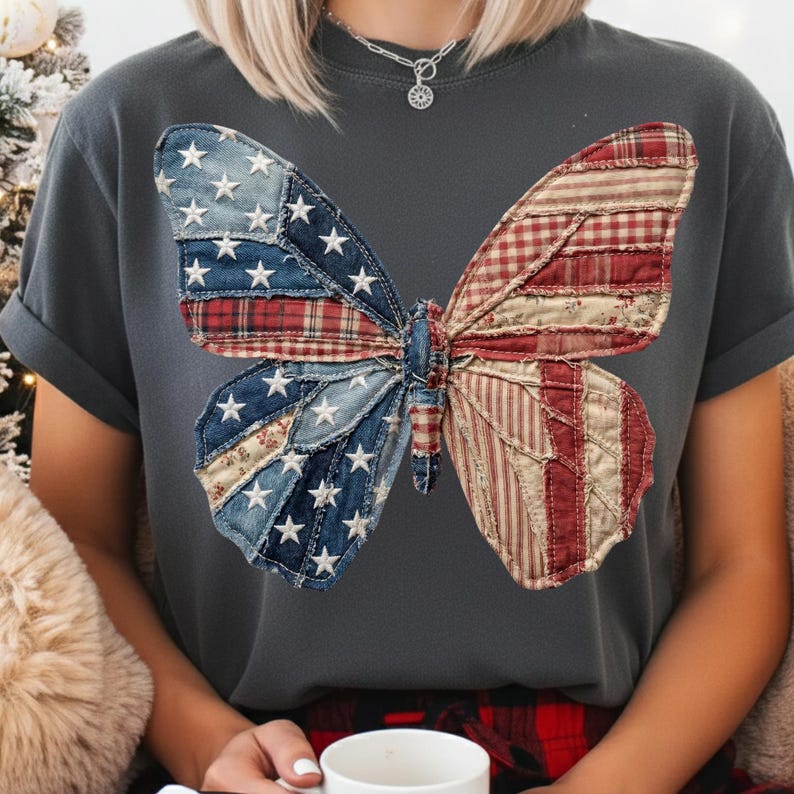 May include: Dark grey t-shirt with a large butterfly design. The butterfly wings are made from patchwork denim, plaid, and striped fabrics in blue, red, and white, forming an American flag pattern. The shirt has short sleeves.
