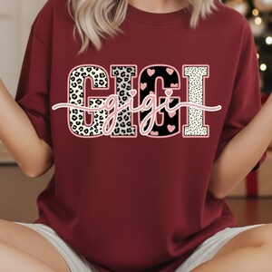 May include: A maroon sweatshirt featuring the word "GIGI" in a bold, multi-textured design. The letters incorporate leopard print, black with pink hearts, and a dotted pattern. The word "gigi" is written in a cursive font across the letters.