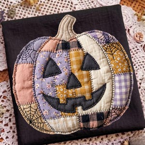 May include: A quilted fabric pumpkin decoration featuring a patchwork design of various patterns including floral prints, gingham, plaid, and spiderwebs in shades of purple, yellow, cream, and black, with dark gray felt eyes and a jagged mouth.