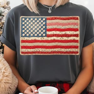May include: Dark grey t-shirt with a fabric American flag design. The flag features a blue denim field with white star accents and red and white striped sections. The shirt has rolled-up sleeves and a relaxed fit.