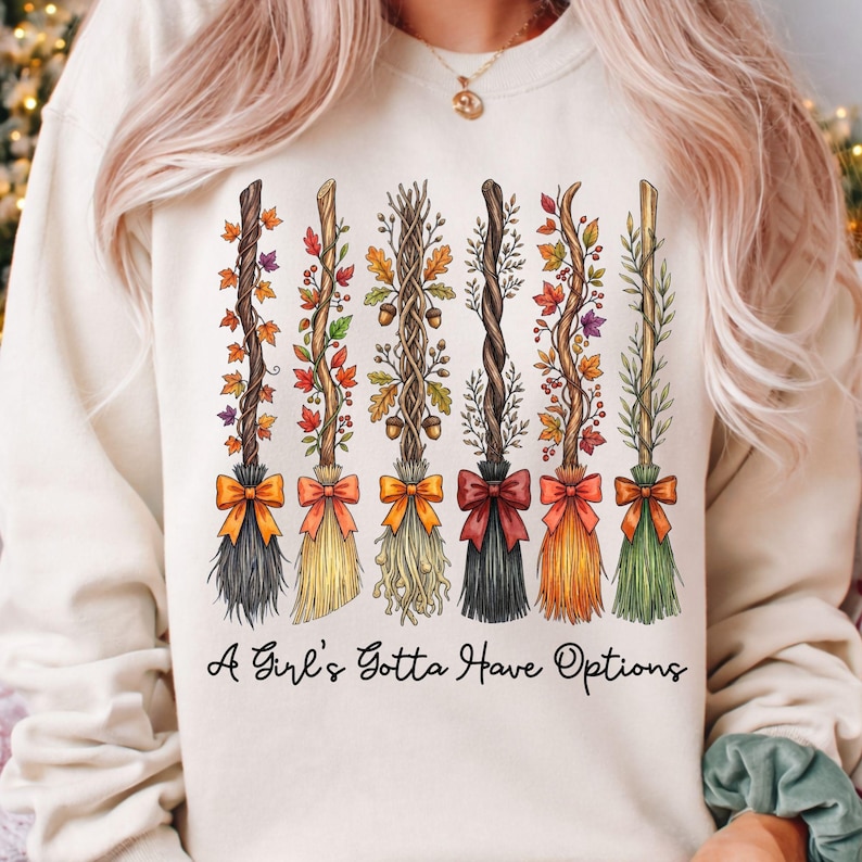 May include: Cream-colored sweatshirt featuring a graphic of six decorative witch brooms. Each broom is adorned with autumn leaves, acorns, and bows in various colors. The text "A Girl's Gotta Have Options" is printed below the brooms.