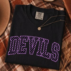 Puede incluir: Una camiseta negra de manga corta con la palabra DEVILS en letras grandes de estilo bloque de color morado con un patrón de puntos negros. La camiseta tiene un cuello redondo y una etiqueta de la marca Comfort Colors visible en el cuello.