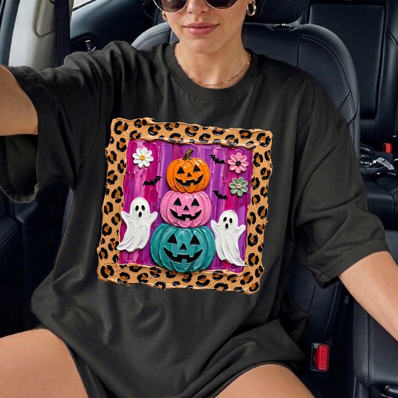 Puede incluir: Una camiseta negra con un gr&aacute;fico cuadrado con borde de estampado de leopardo. El centro muestra una pila de tres calabazas de Halloween en naranja, rosa y turquesa, flanqueadas por dos fantasmas blancos, peque&ntilde;os murci&eacute;lagos negros y tres flores peque&ntilde;as sobre un fondo morado.