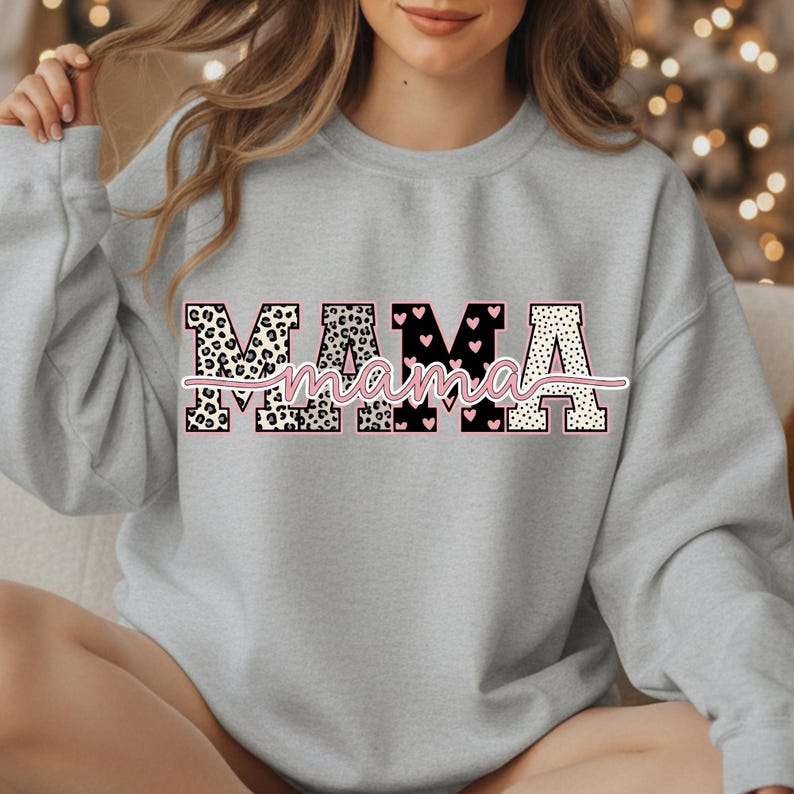 May include: A light gray sweatshirt with the word "MAMA" in a decorative font. The letters have a mix of leopard print, black hearts, and polka dots. The word "mama" is written in a cursive font in pink.