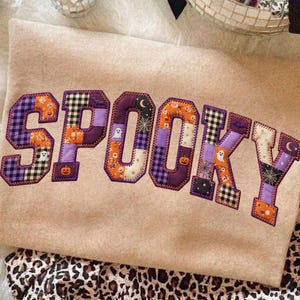 May include: A beige sweatshirt featuring the word SPOOKY in large, block-style letters. Each letter is constructed from various fabric patches in purple, orange, black, and white, displaying patterns of pumpkins, ghosts, bats, and spiderwebs.