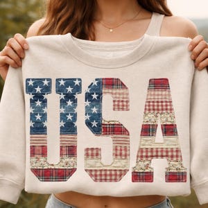 May include: Cream-coloured sweatshirt featuring the word "USA" in a patchwork design. The letters are decorated with denim, stars, and red plaid patterns, creating a patriotic theme.