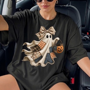 Op de afbeelding: Een zwart T-shirt met korte mouwen en een afbeelding van een geest gemaakt van verschillende stoffen lapjes. De geest draagt een grote strik met patroon op het hoofd en draagt een klein oranje pompoenemmertje. Het ontwerp bevat ruitjes en bloemmotieven.