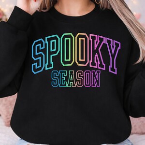 May include: A black long-sleeve sweatshirt featuring the text SPOOKY SEASON printed in a vibrant rainbow gradient. The lettering is styled in a bold, blocky varsity font with a thin outline, centered prominently on the chest of the garment.