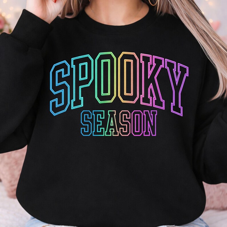 May include: A black long-sleeve sweatshirt featuring the text SPOOKY SEASON printed in a vibrant rainbow gradient. The lettering is styled in a bold, blocky varsity font with a thin outline, centered prominently on the chest of the garment.