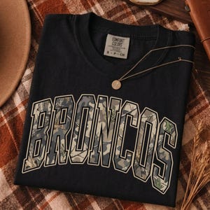 Puede incluir: Una camiseta negra de manga corta con la palabra BRONCOS en una fuente universitaria grande y arqueada. Las letras están rellenas con un patrón de camuflaje en tonos verdes, marrones y grises. La camiseta tiene una etiqueta Comfort Colors en el cuello.
