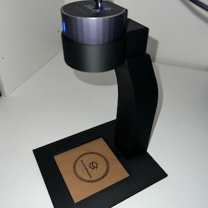 May include: A black 3D printed stand with a circular base and a curved arm. A small, gray, cylindrical speaker is mounted on the arm. The speaker has a blue light on the side. The stand has a brown cardboard square with a logo on it.