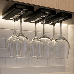 May include: A black wine glass rack installed under a cabinet, holding six clear wine glasses. The glasses are suspended upside down, showcasing their elegant design. The background features a white tiled backsplash and a light-colored countertop.