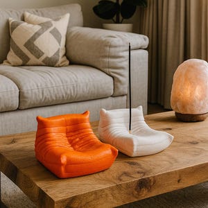 Peut inclure: Deux porte-encens décoratifs en forme de petits fauteuils. L'un est orange vif, l'autre blanc cassé. Un bâton d'encens est placé dans le support blanc. Ils sont posés sur une table basse en bois dans un salon.