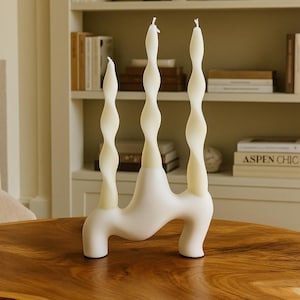 Könnte beinhalten: Ein weißer, skulpturaler Kerzenhalter mit drei gedrehten Kerzen. Die Kerzen sind cremefarben und sitzen auf einem modernen, abstrakten Sockel. Der Kerzenhalter steht auf einem Holztisch.