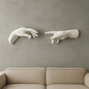 Op de afbeelding: Witte wandkunst met twee handen in een aanrakingsgebaar, gemonteerd op een grijze muur. De handen zijn realistisch gebeeldhouwd, met gedetailleerde vingers en handpalmen. Een beige bank staat eronder.