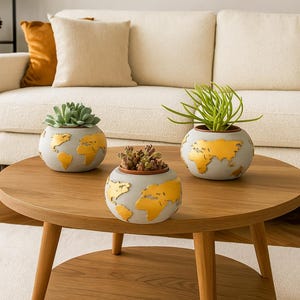 May include: Three gray globe-shaped planters with gold continents, each holding different succulents, sit on a round wooden coffee table. The planters are a decorative home decor item. The table is in front of a cream-colored couch.