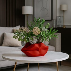 May include: A red ceramic lip-shaped vase filled with a variety of green and dried flowers sits on a white marble-topped coffee table with gold legs. The vase is the focal point, with a neutral-toned sofa and artwork in the background.