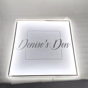 May include: A square, illuminated sign displaying the text "Denise's Den" in a cursive font. The sign has a white background with a black border and a square outline around the text. The sign is lit from within.